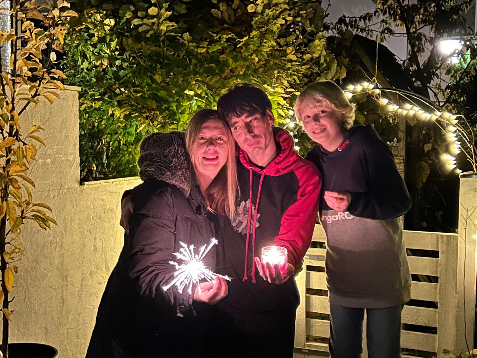 Drei Menschen stehen abends dicht beieinander in einem Garten unter einer Lichterkette: links eine Frau mit einer brennenden Wunderkerze, in der Mitte ein Mann im rot-schwarzen Kapuzenpullover mit einem Windlicht in der Hand, rechts ein Jugendlicher.